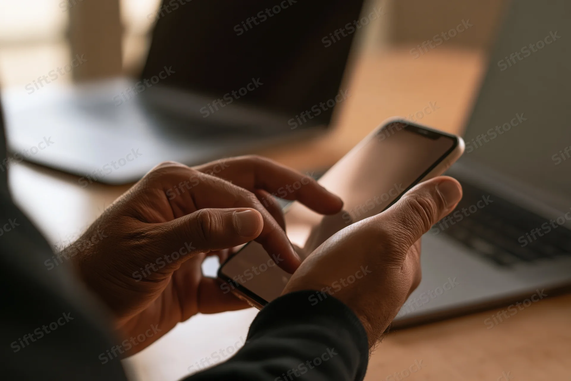 Man tapping smartphone with both hands at desk with laptop in background