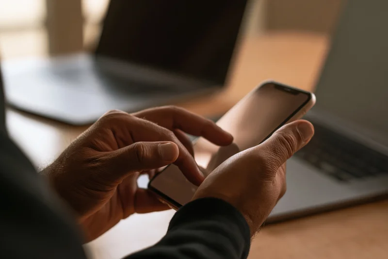 Man tapping smartphone with both hands at desk with laptop in background