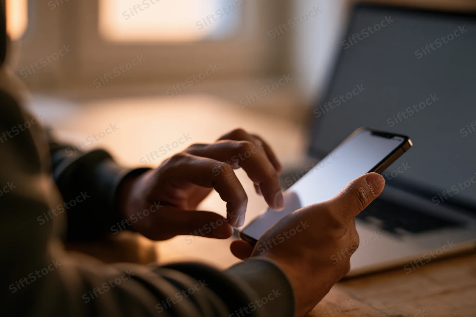 Man tapping on smartphone at desk with laptop nearby