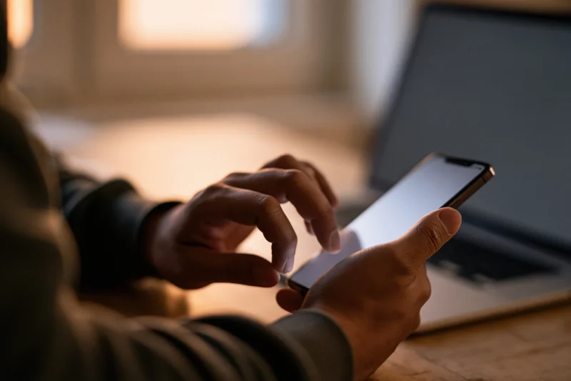 Man tapping on smartphone at desk with laptop nearby