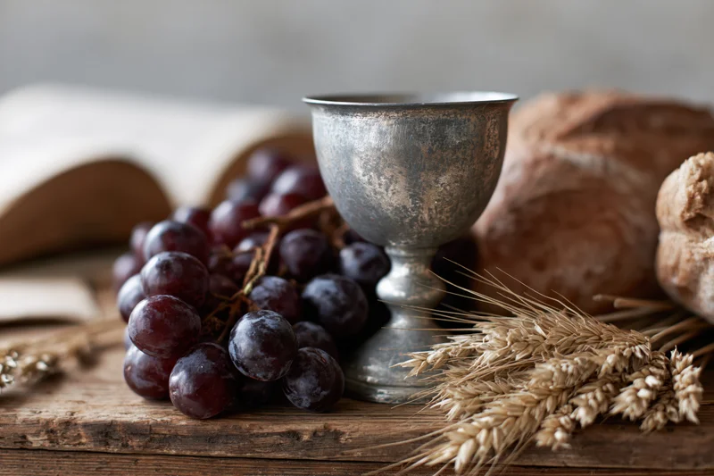 Pewter communion chalice with dark red grapes, bread, and wheat stalks on a rustic wooden table