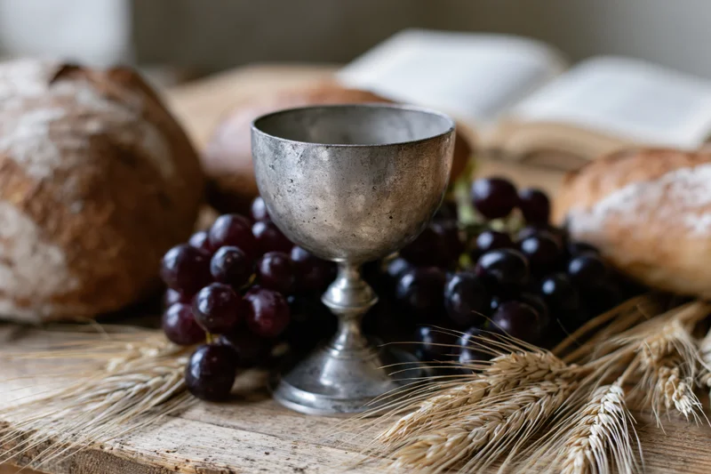 Pewter chalice surrounded by dark grapes and loaves of bread on a wooden board with wheat stalks