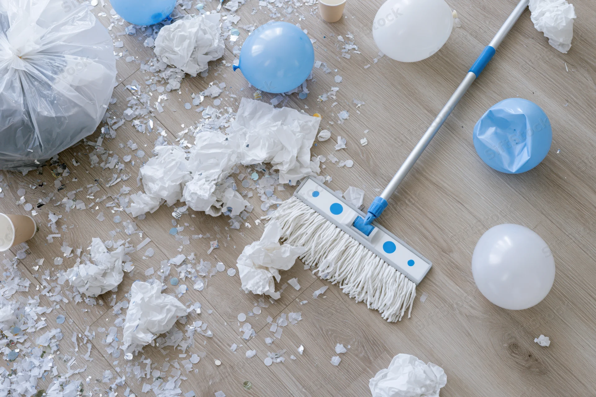 Overhead view of post-party mess with balloons, confetti and trash on light wood laminate floor and cleaning mop nearby