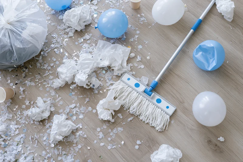 Overhead view of post-party mess with balloons, confetti and trash on light wood laminate floor and cleaning mop nearby