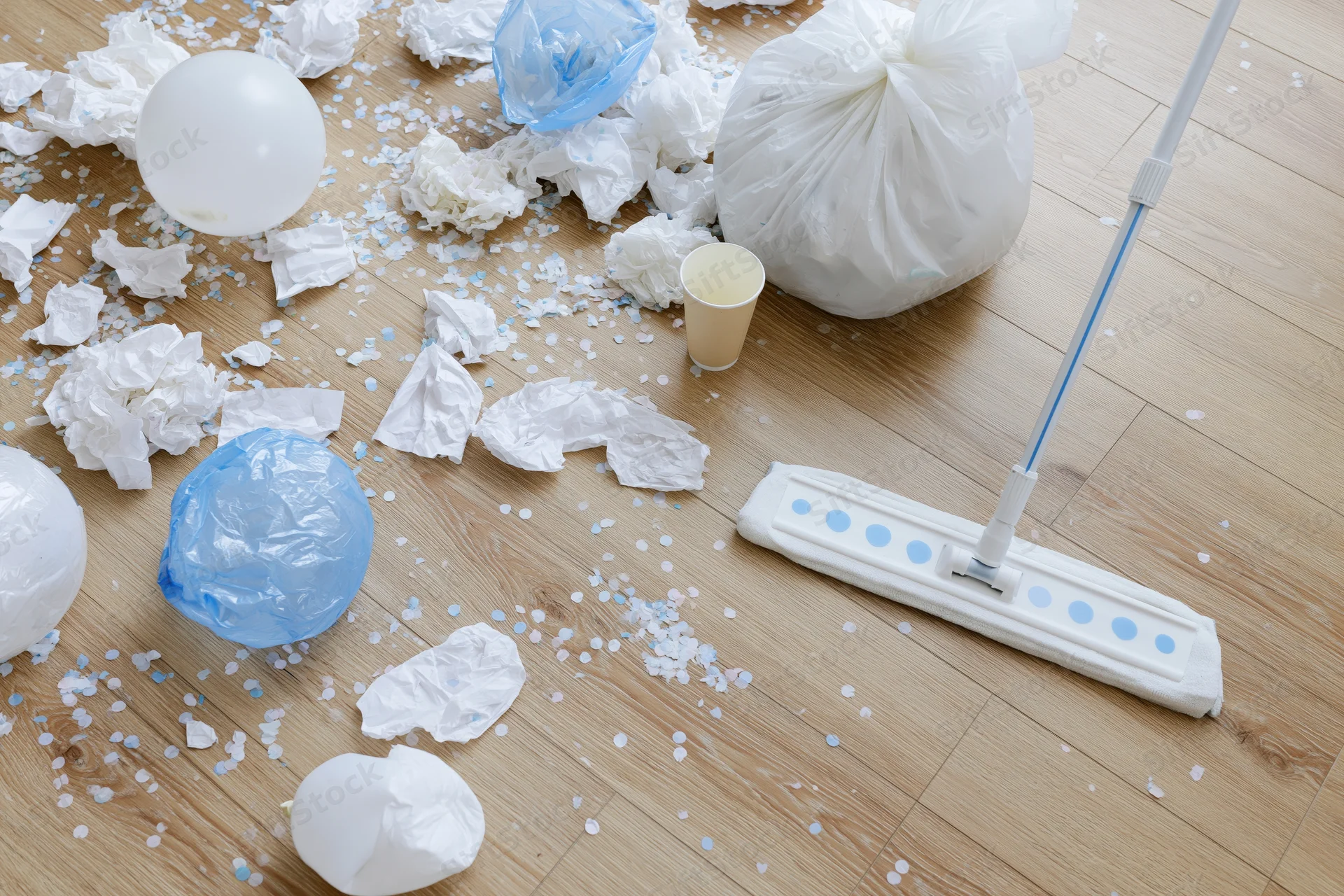 Post-party mess scattered across light wood laminate floor with mop and trash bag present