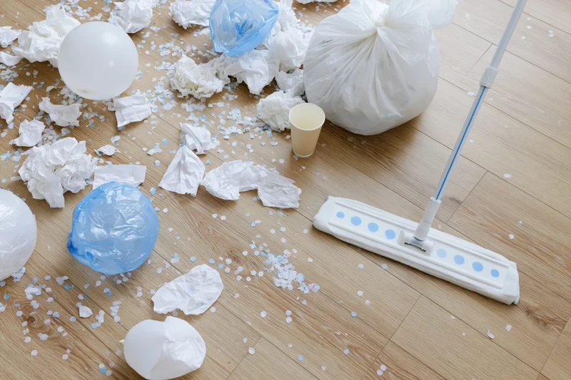 Post-party mess scattered across light wood laminate floor with mop and trash bag present