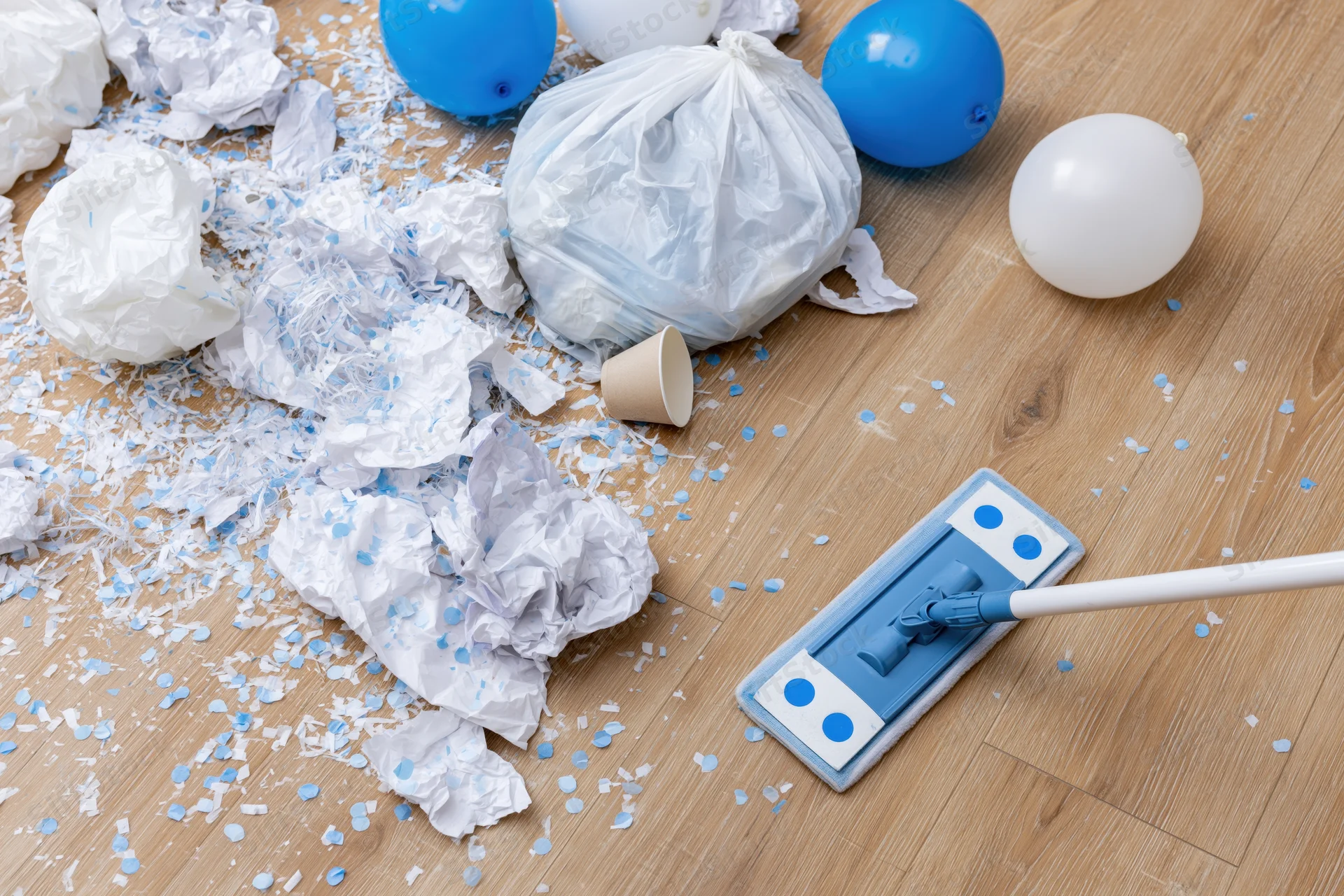 Post-party mess with balloons, confetti and trash on a light wood laminate floor, mop nearby