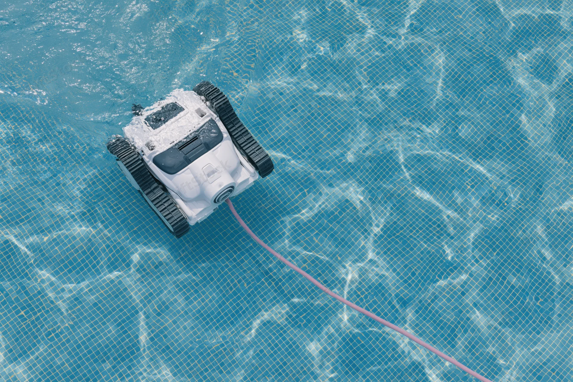 Robotic pool cleaner with black rubber tracks and white plastic body moving across tiled pool floor, connected by cord