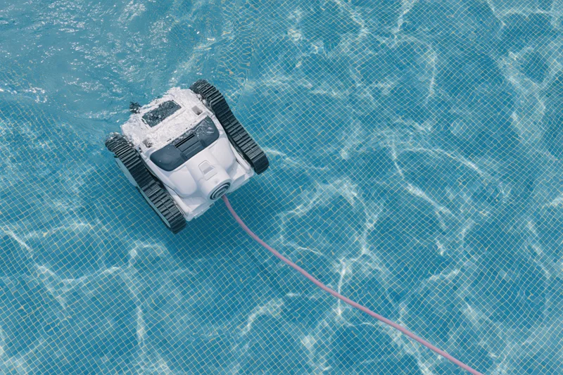 Robotic pool cleaner with black rubber tracks and white plastic body moving across tiled pool floor, connected by cord