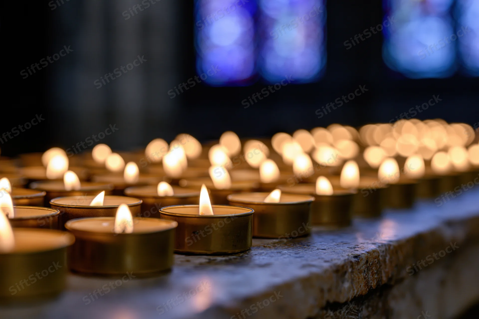 Rows of burning votive candles in metal holders on a stone surface inside a church with stained glass windows in the background
