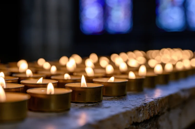 Rows of burning votive candles in metal holders on a stone surface inside a church with stained glass windows in the background