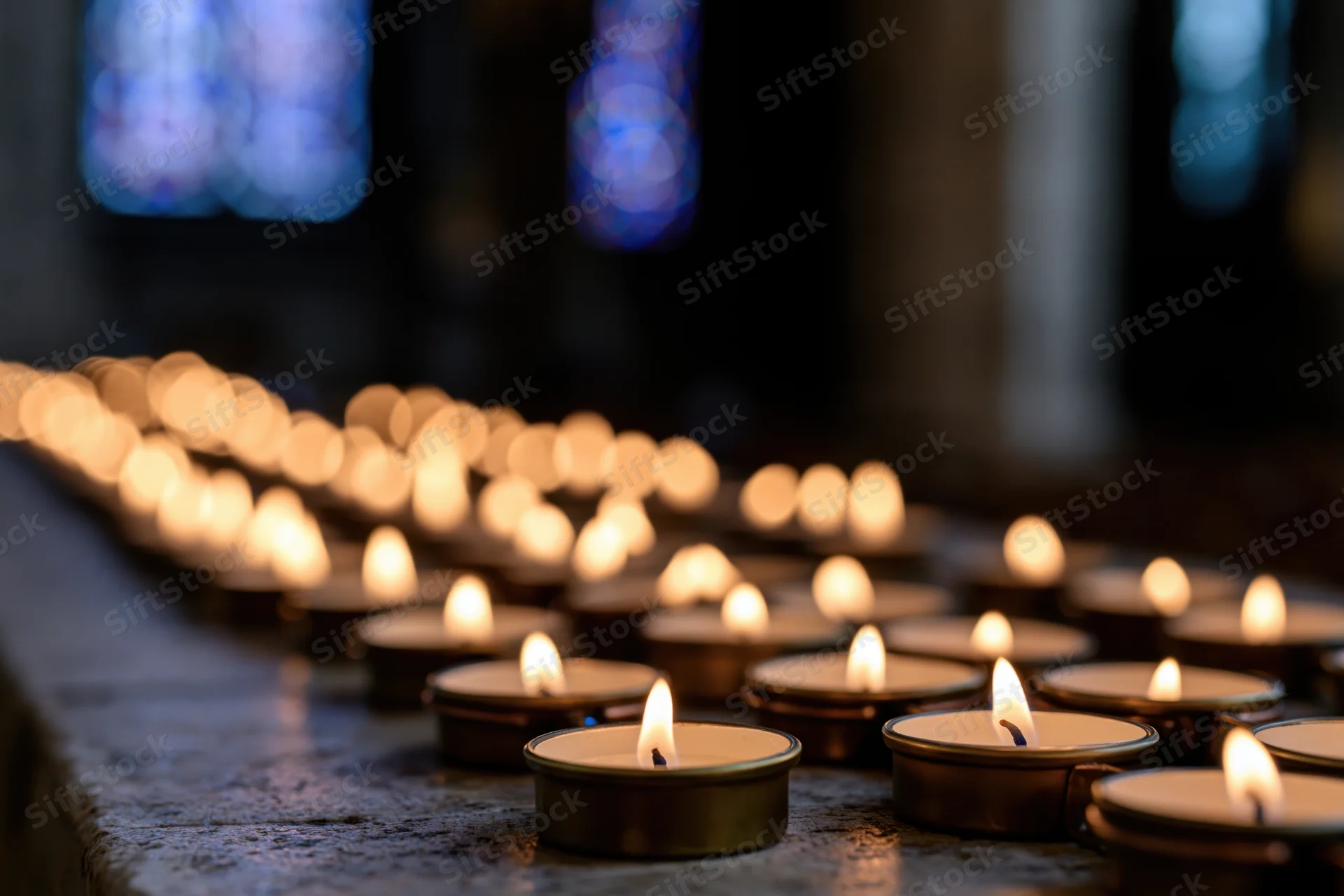 Rows of lit votive candles in metal holders arranged on a stone surface inside a church with stained glass windows in the background
