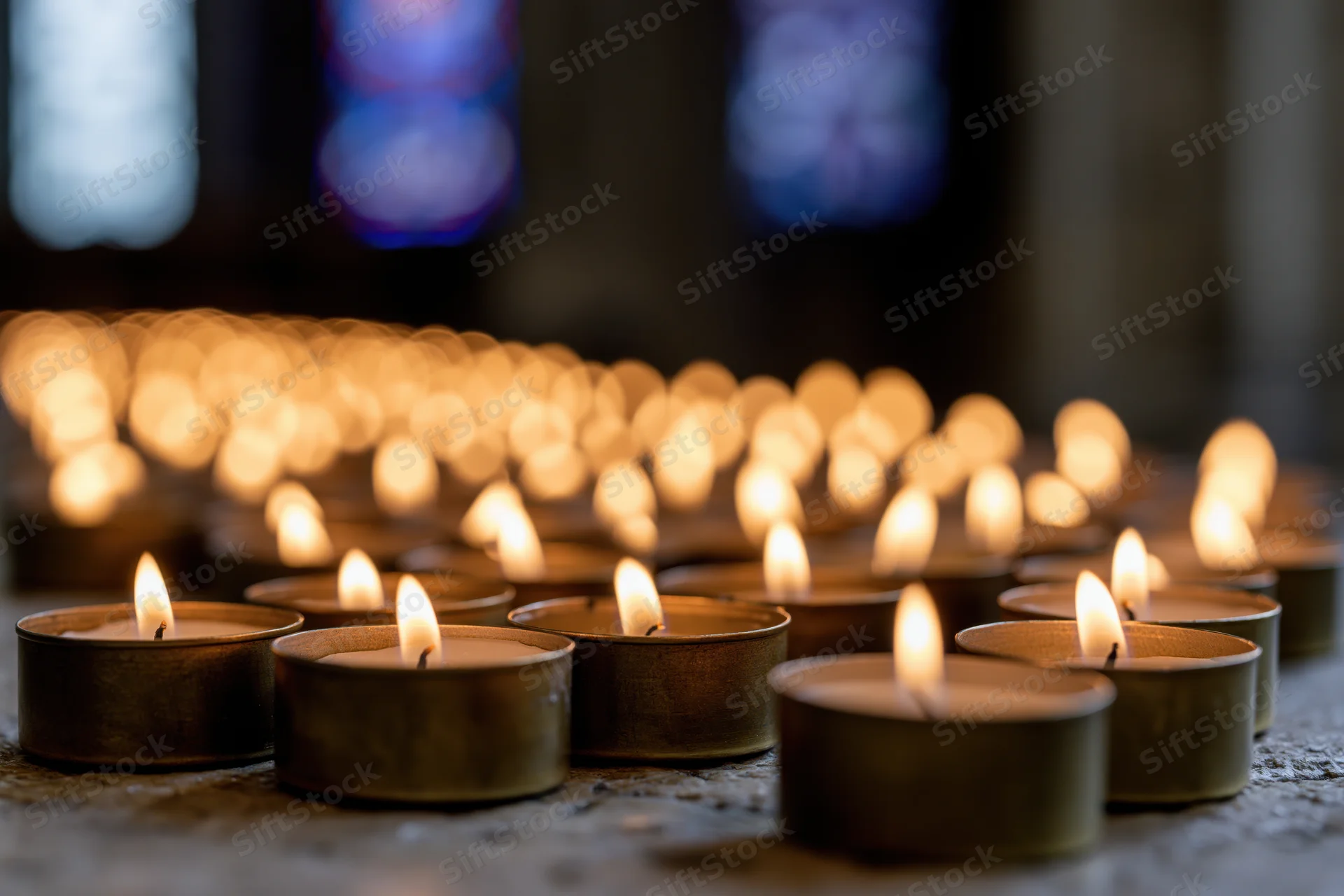 Rows of lit votive candles in metal holders on stone floor inside church with stained glass windows in background