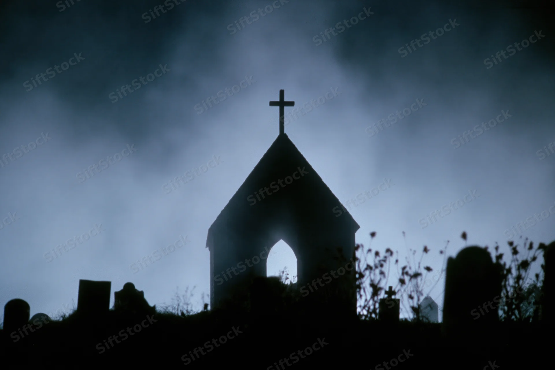 Silhouette of a gothic chapel with a cross on its peak in a foggy graveyard at night