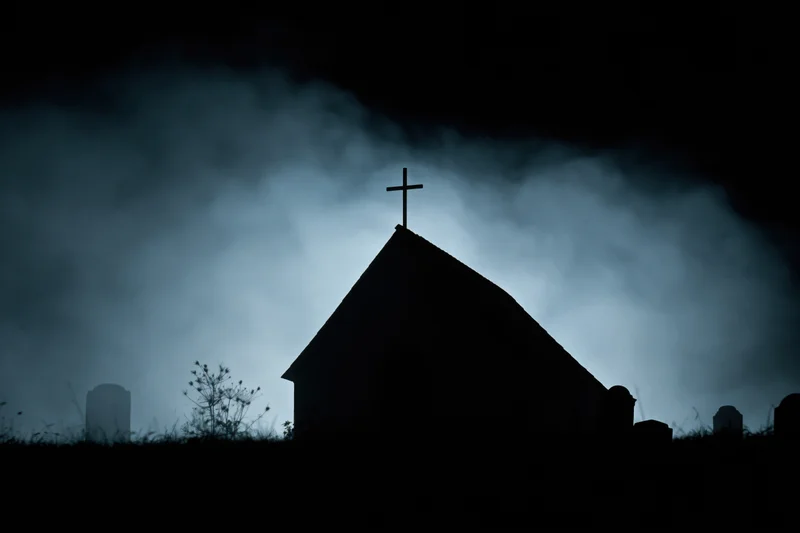 Silhouette of a gothic chapel with a cross on its peak surrounded by fog and tombstones at night