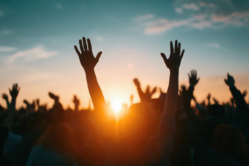 Silhouetted crowd raising hands toward sunset at outdoor concert