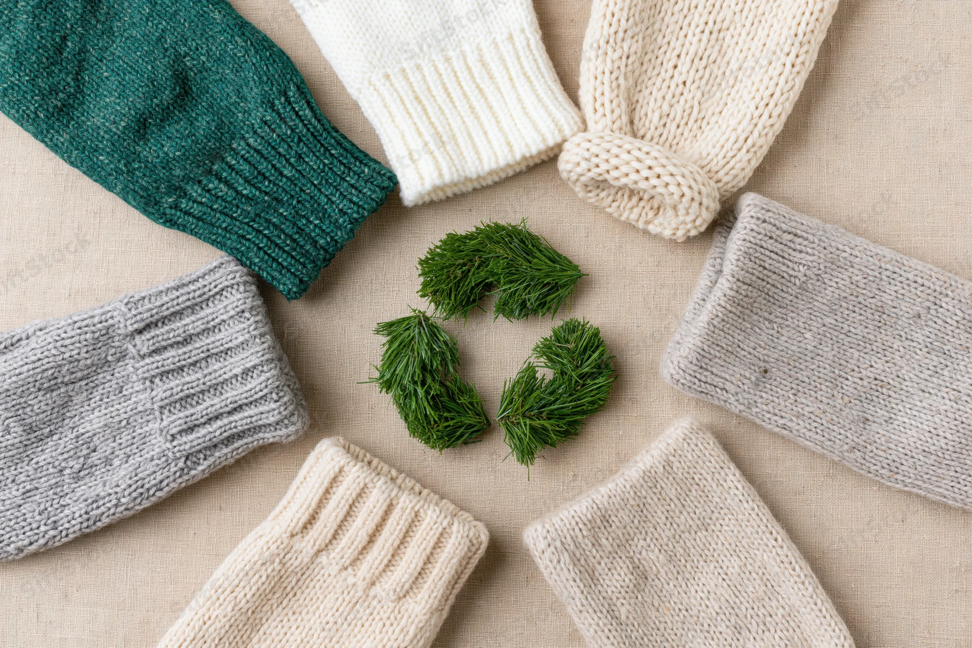 Knitted sweater sleeves arranged in a circle around a pine needle recycling symbol on a neutral fabric background