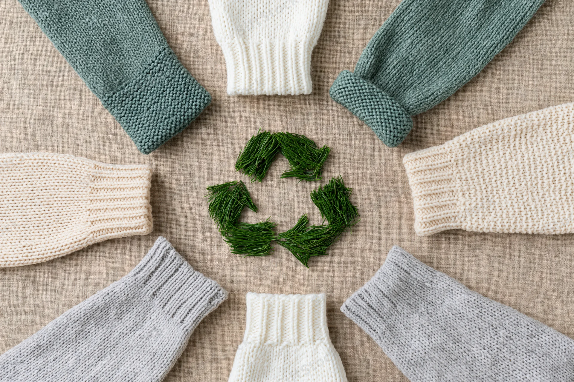 Knitted sweater sleeves arranged in a circle around a green recycling symbol made of sprigs on a beige fabric background