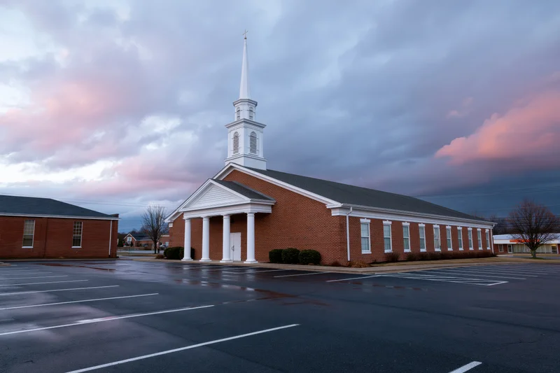 Brick baptist church with white steeple and columns in front of wet parking lot under cloudy twilight sky