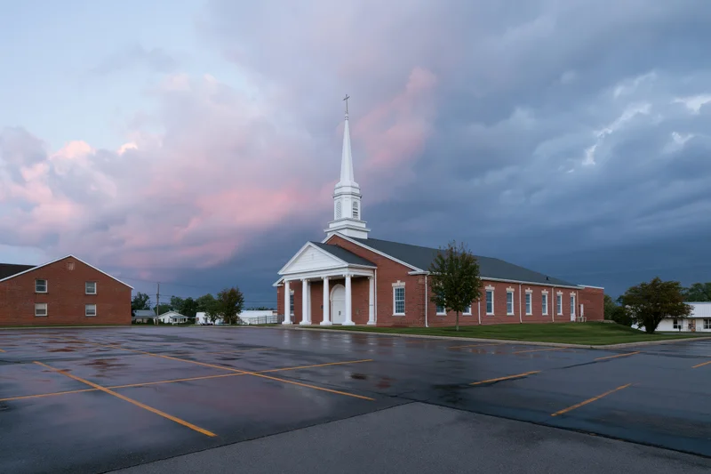 Brick baptist church with white steeple and cross under cloudy twilight sky, viewed from wet parking lot