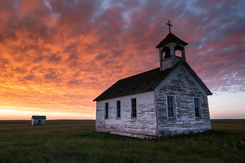 Small abandoned white clapboard church with bell tower standing in a grassy prairie field at sunset