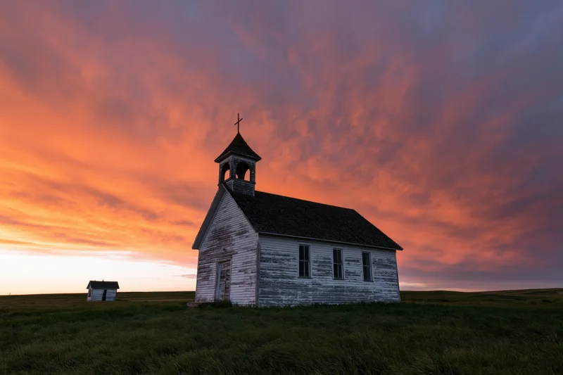 Small abandoned white clapboard church with bell steeple and cross at sunset on prairie field