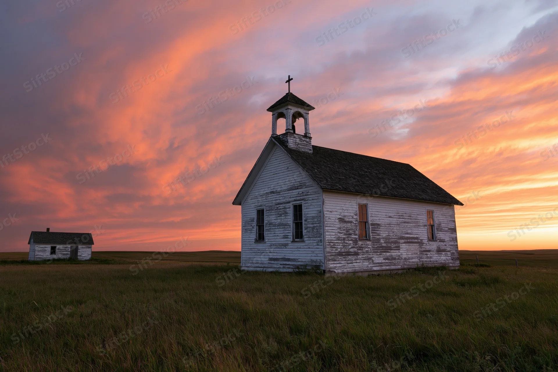 Abandoned white clapboard church with bell steeple and cross standing in a grassy prairie field at sunset