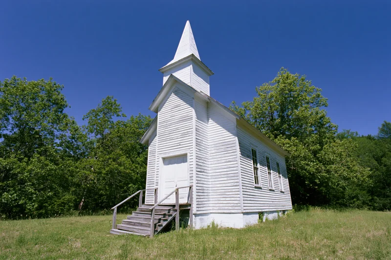 Small white wooden church with steeple and steps in a grassy field surrounded by trees under a clear blue sky
