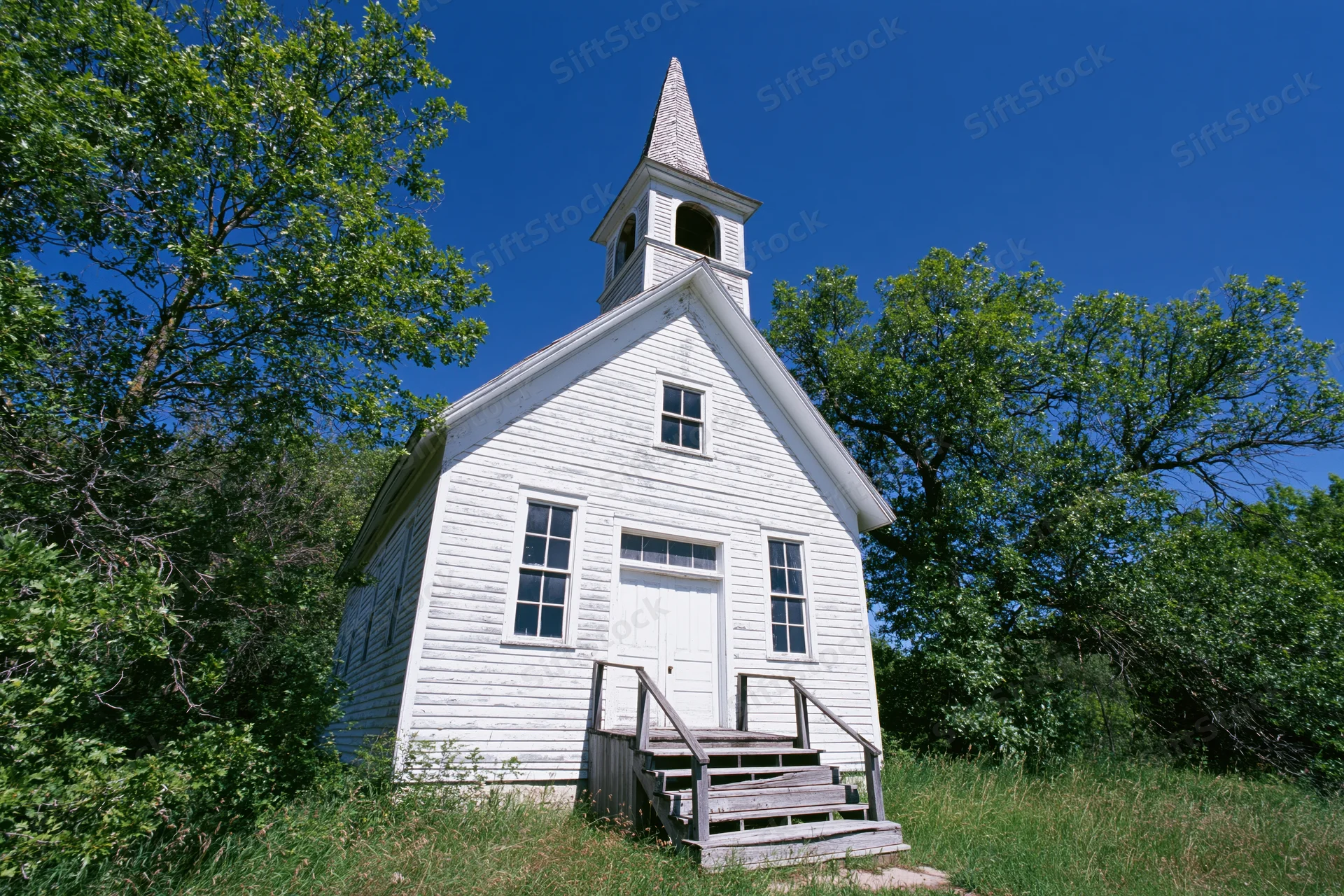 Small white wooden church with steeple and steps surrounded by trees and grass under clear blue sky