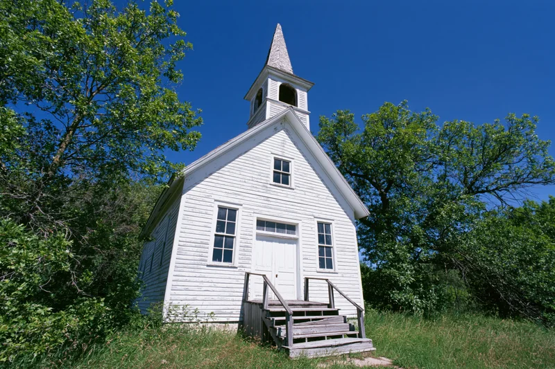 Small white wooden church with steeple and steps surrounded by trees and grass under clear blue sky
