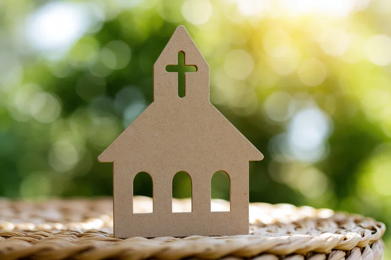 Cardboard church cutout standing on a woven wicker surface with green foliage and sunlight in the background