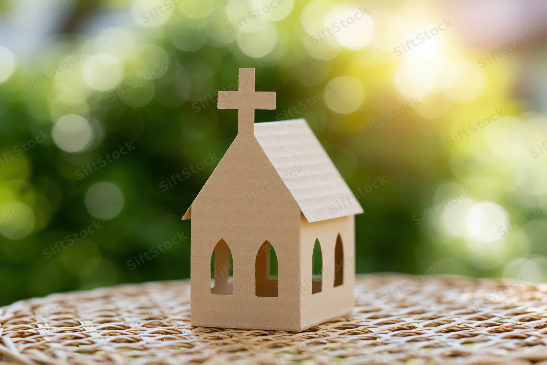 Small cardboard church model standing on a woven mat with blurred green foliage and sunlight in the background