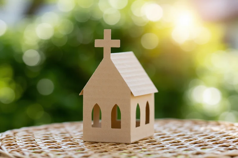 Small cardboard church model standing on a woven mat with blurred green foliage and sunlight in the background