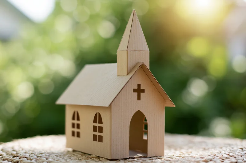 Small cardboard church model with cross and steeple on woven mat outdoors