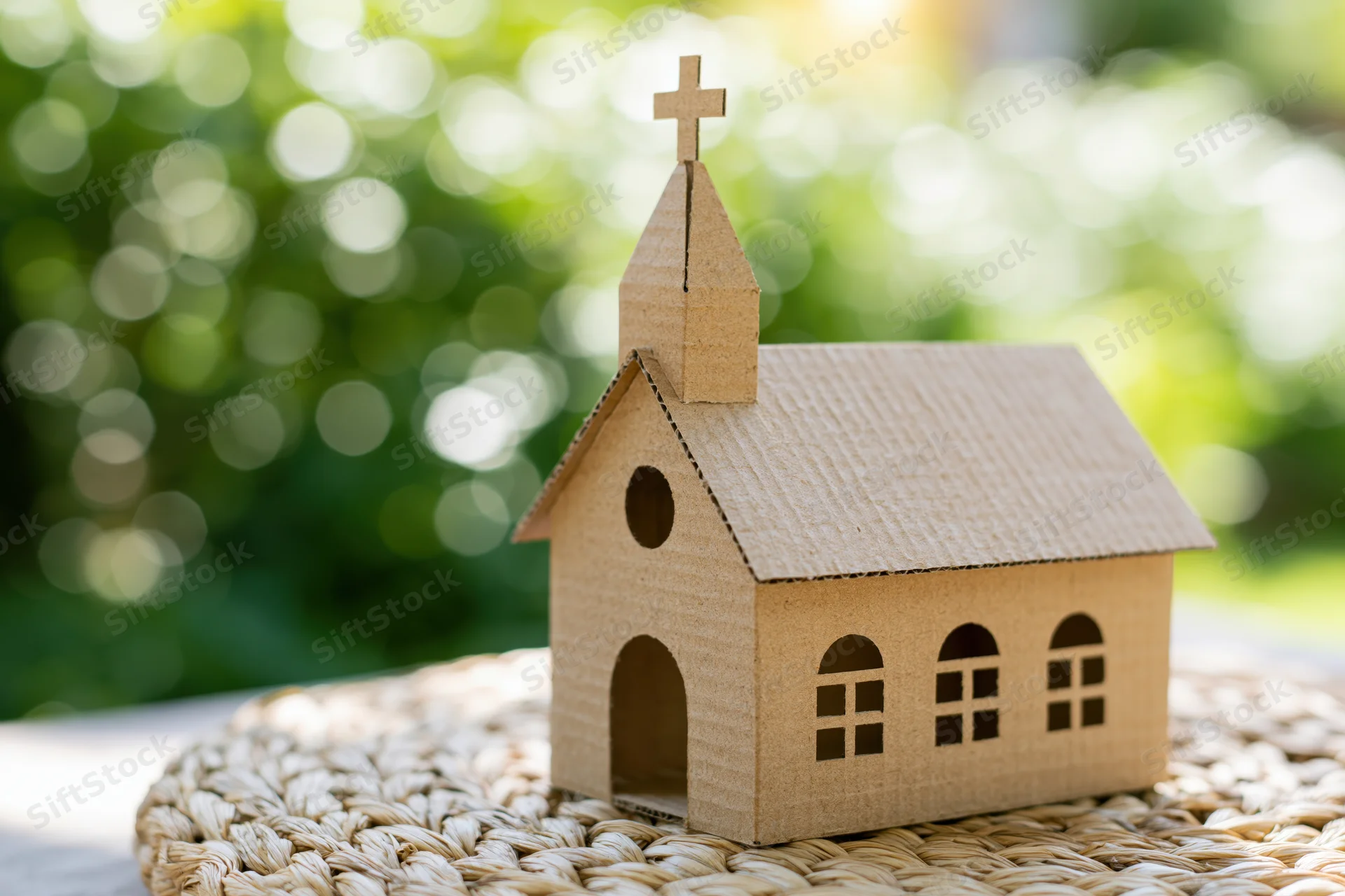 Small cardboard church model with cross on steeple placed on woven mat outdoors