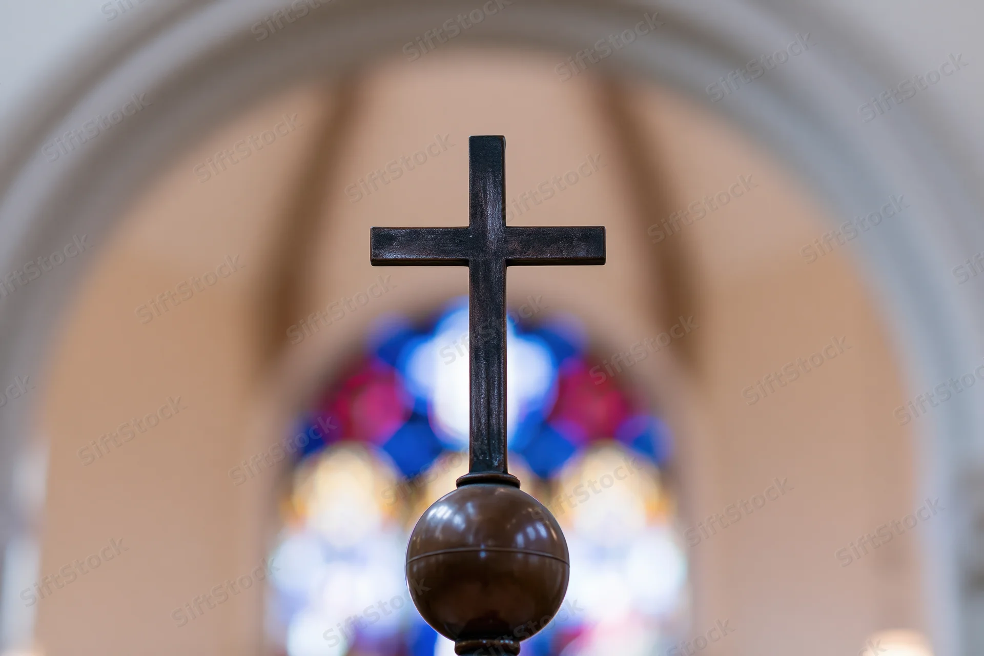 Dark bronze cross mounted on a sphere centered in frame with stained glass window in background through an arch
