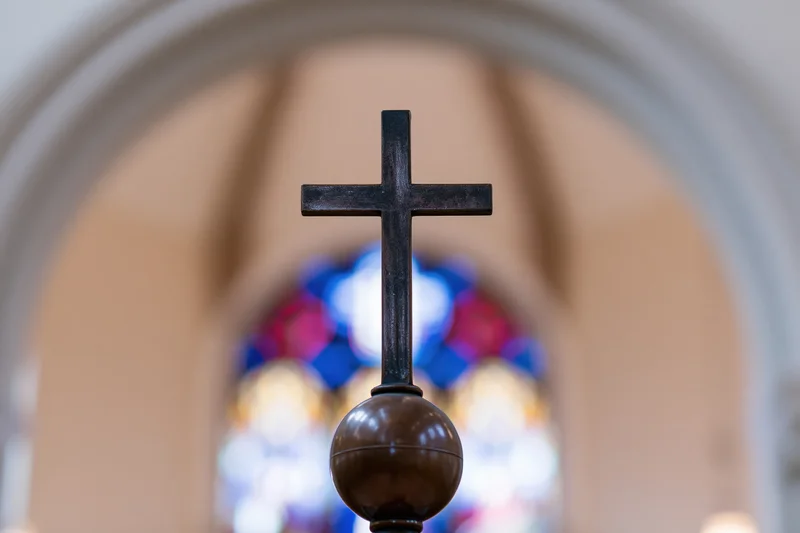 Dark bronze cross mounted on a sphere centered in frame with stained glass window in background through an arch