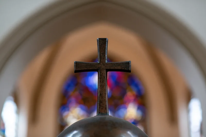 Bronze cross mounted on a sphere centered in a church interior with stained glass window in background