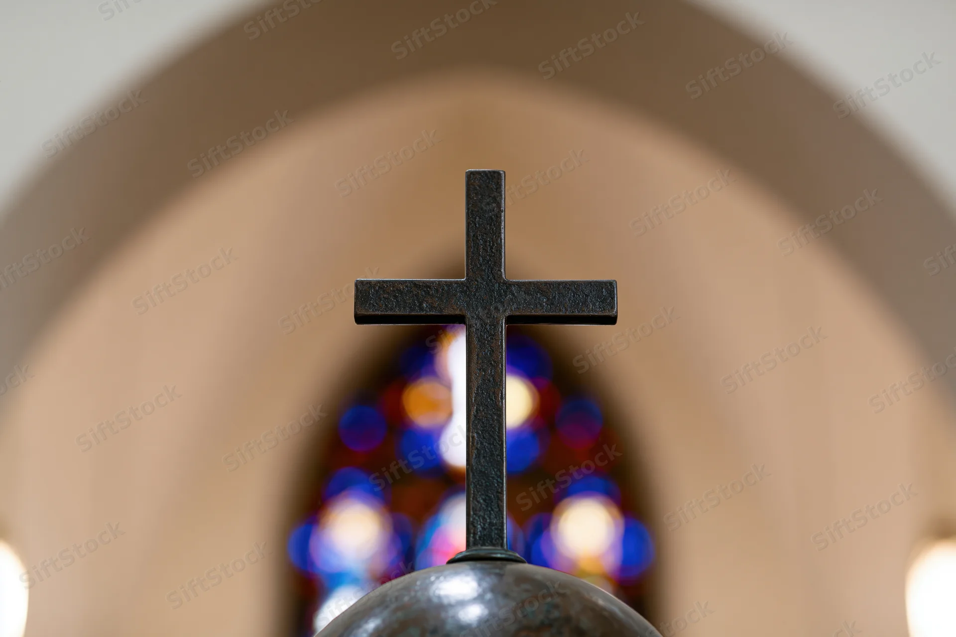 Dark bronze cross mounted on a sphere centered in frame with stained glass bokeh in background