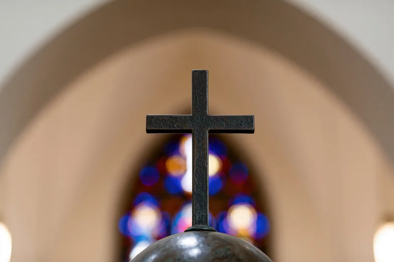 Dark bronze cross mounted on a sphere centered in frame with stained glass bokeh in background