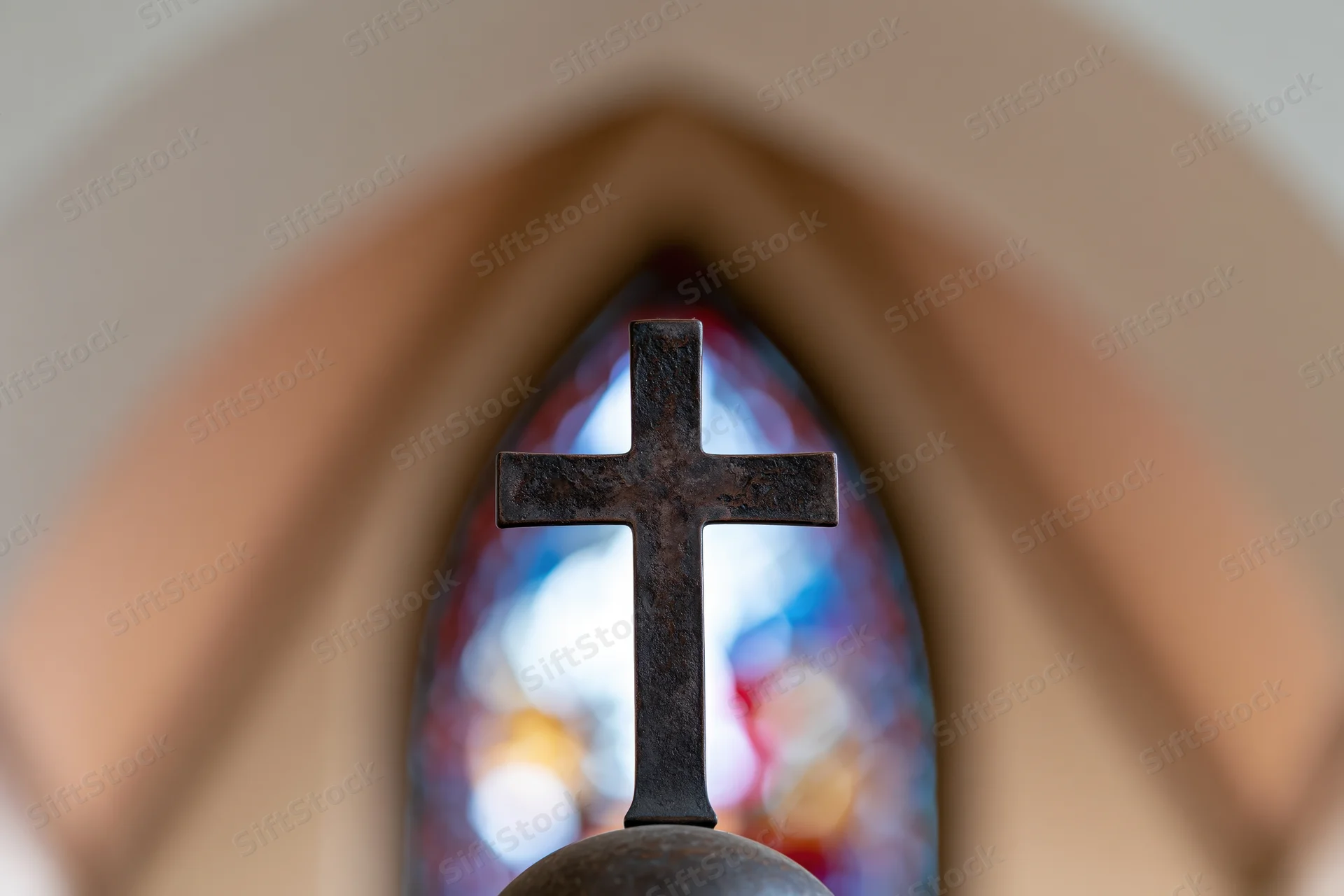 Dark bronze cross mounted on sphere centered in frame with stained glass window in background