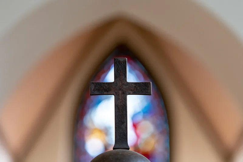 Dark bronze cross mounted on sphere centered in frame with stained glass window in background