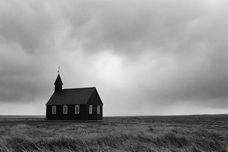Small dark wooden church with steeple standing alone on open grassy field under cloudy sky