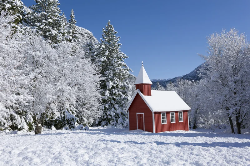 Small red wooden chapel with pointed steeple surrounded by snow-covered trees and mountains