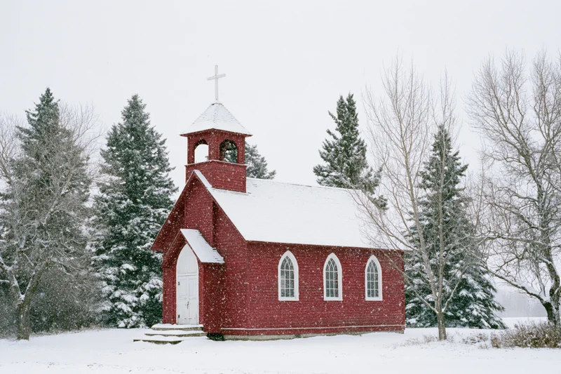 Small red wooden country church with bell tower and white cross in snowfall surrounded by pine trees