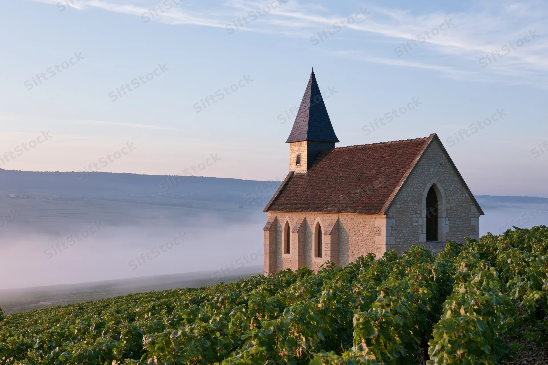Small stone chapel with dark slate steeple perched atop a hill overlooking a foggy valley surrounded by vineyards at dawn