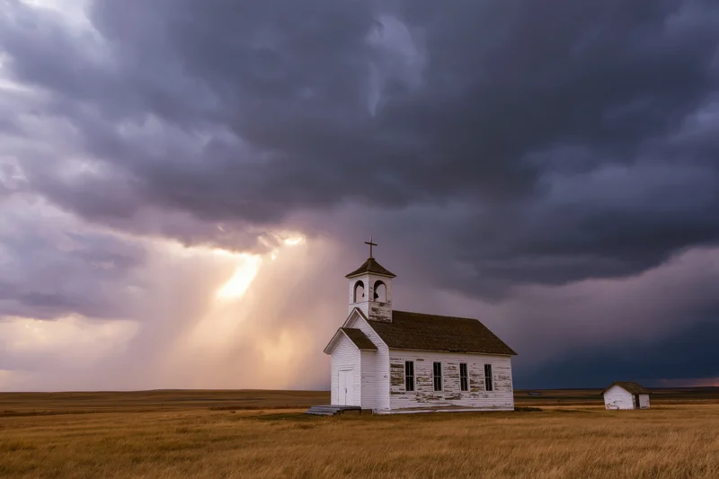 Small white clapboard church with bell and cross standing alone in a grassy prairie field under stormy sky with sunbeams breaking through clouds