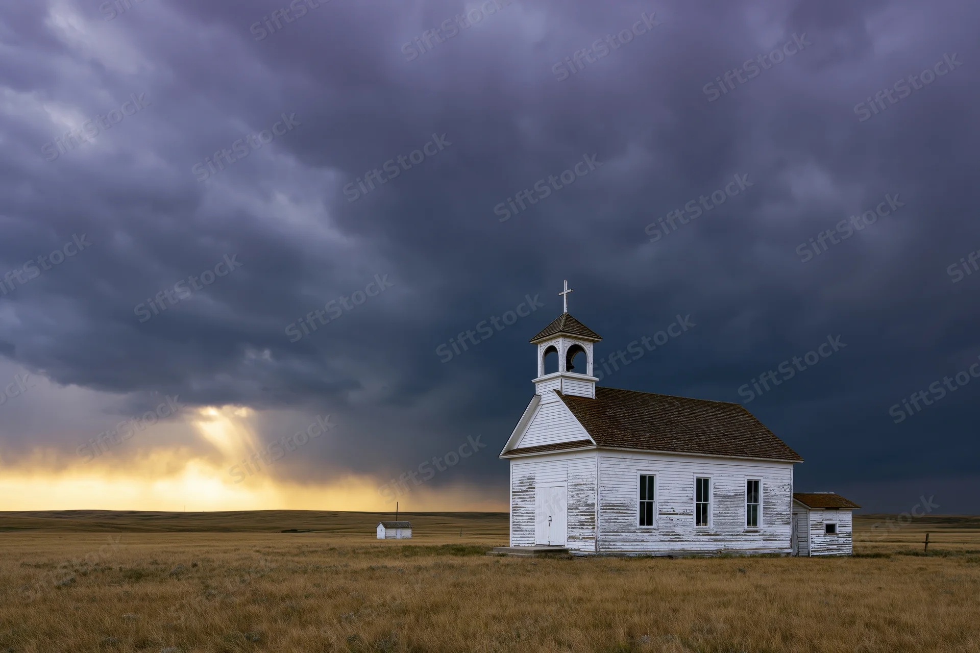 Small white weathered church on prairie under stormy sky with sunbeams breaking through clouds