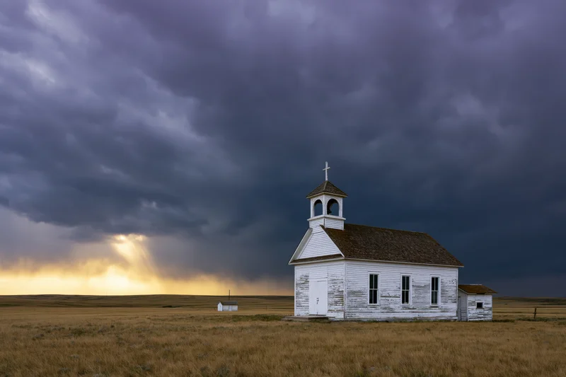 Small white weathered church on prairie under stormy sky with sunbeams breaking through clouds