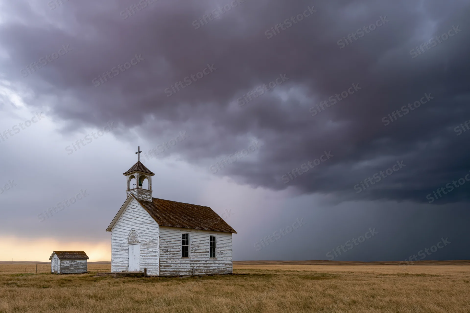 Small weathered white clapboard church with bell tower standing alone in a grassy prairie under stormy dark clouds